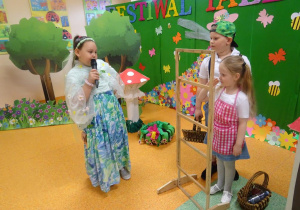 Dziewczynka z mikrofonem w ręku recytuje wiersz zwrócona w kierunku dwójki dzieci.